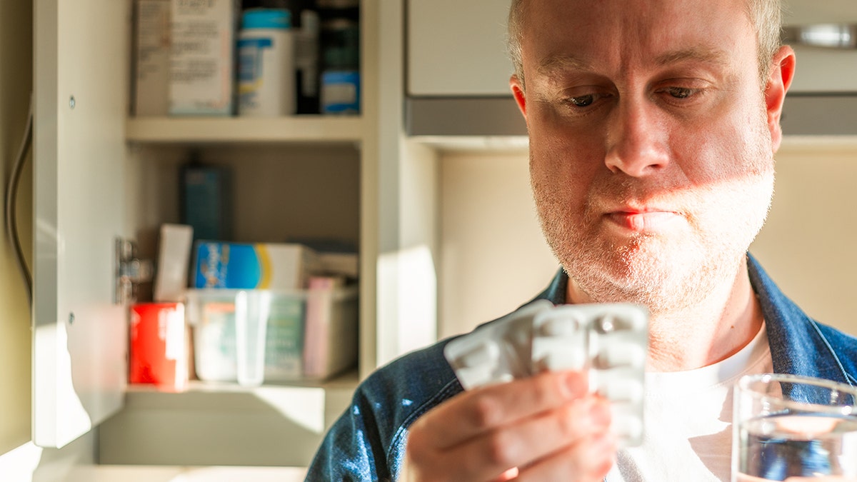 The man looked at a package of painkillers in the kitchen while holding a glass of water.